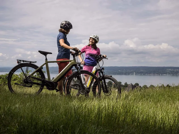 Zwei Radfahrer mit Helmen stehen auf einer Wiese mit Blick auf einen See