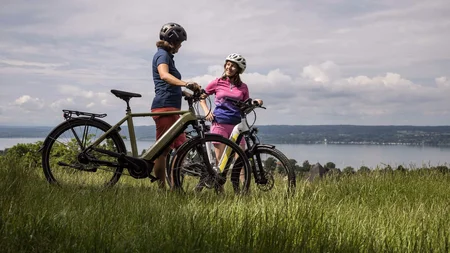 Zwei Radfahrer mit Helmen stehen auf einer Wiese mit Blick auf einen See