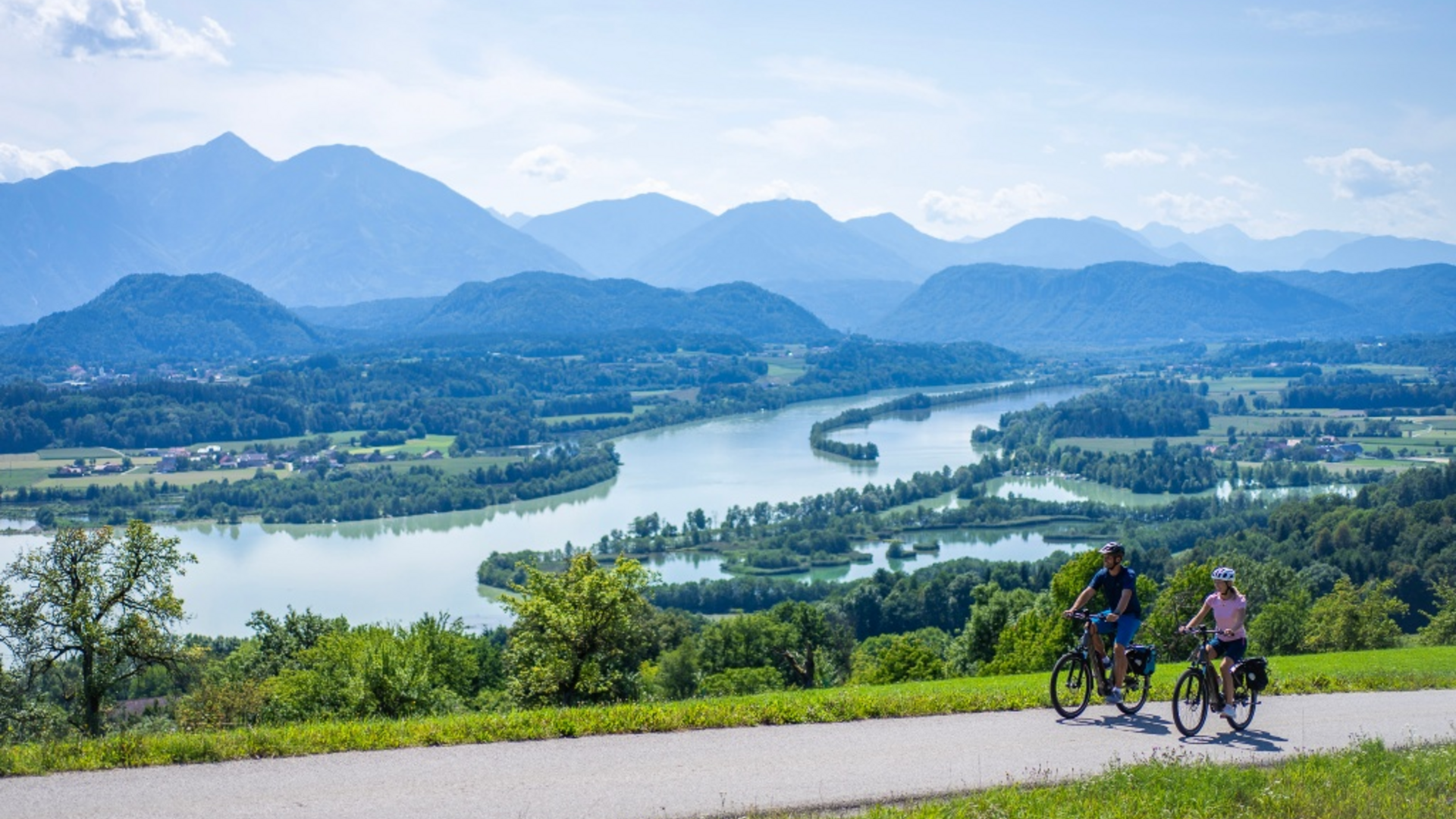 Two cyclists on mountain road with river and mountains in the background