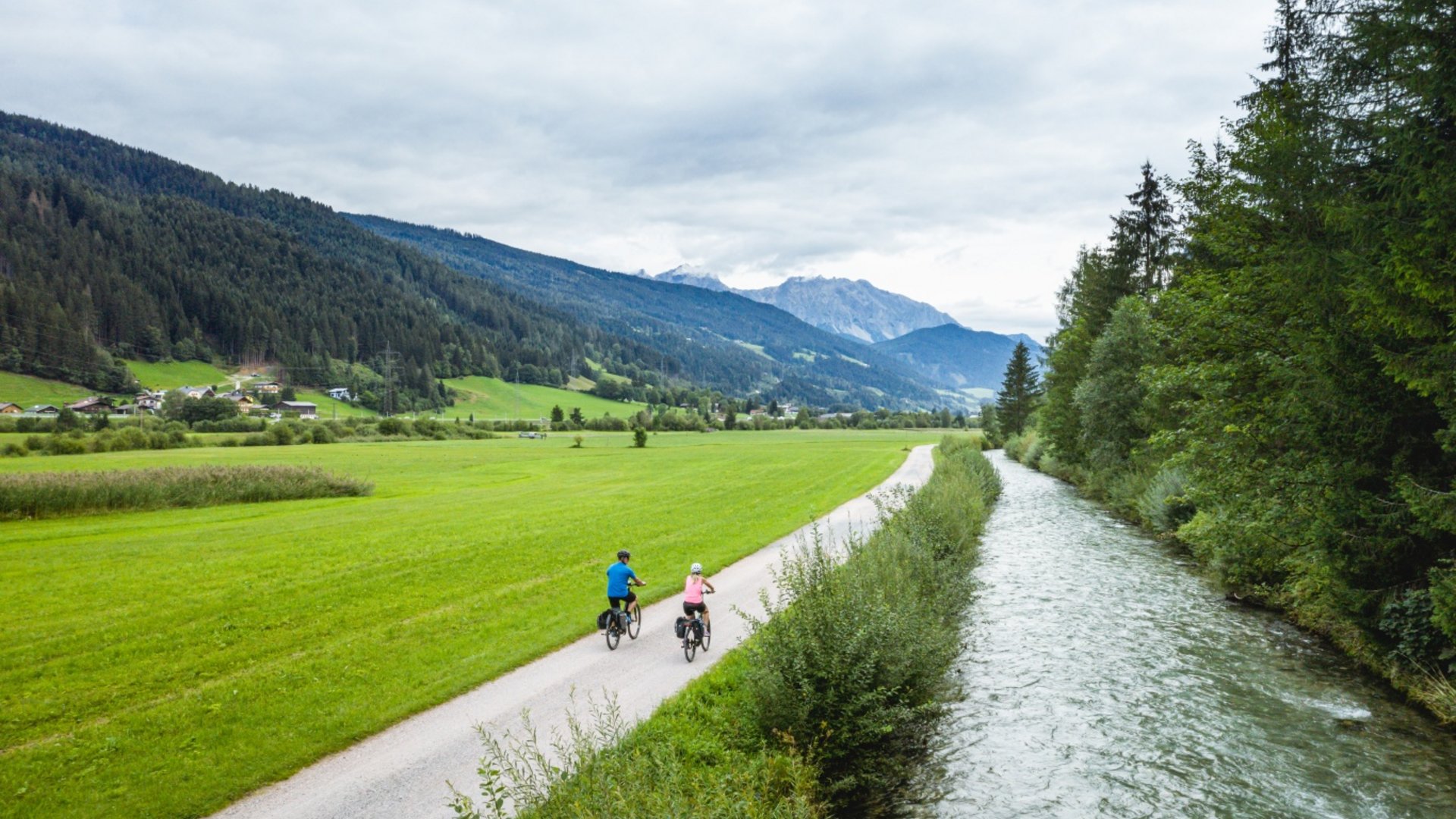 Two cyclists on path beside river in green mountain landscape