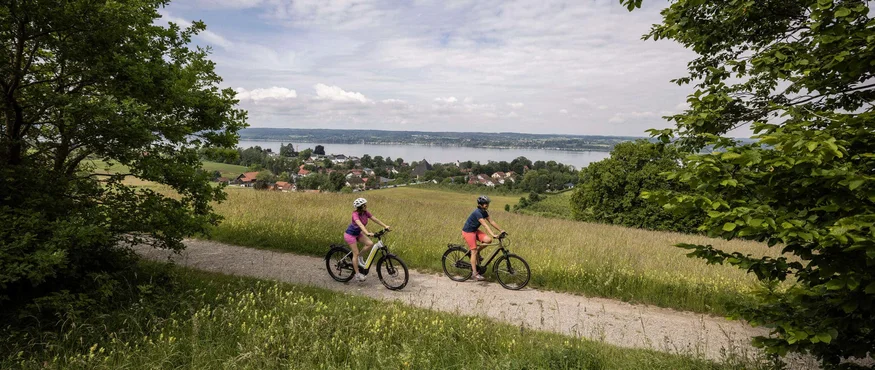 Zwei Personen fahren mit dem Fahrrad auf einem Weg mit Aussicht auf einen See