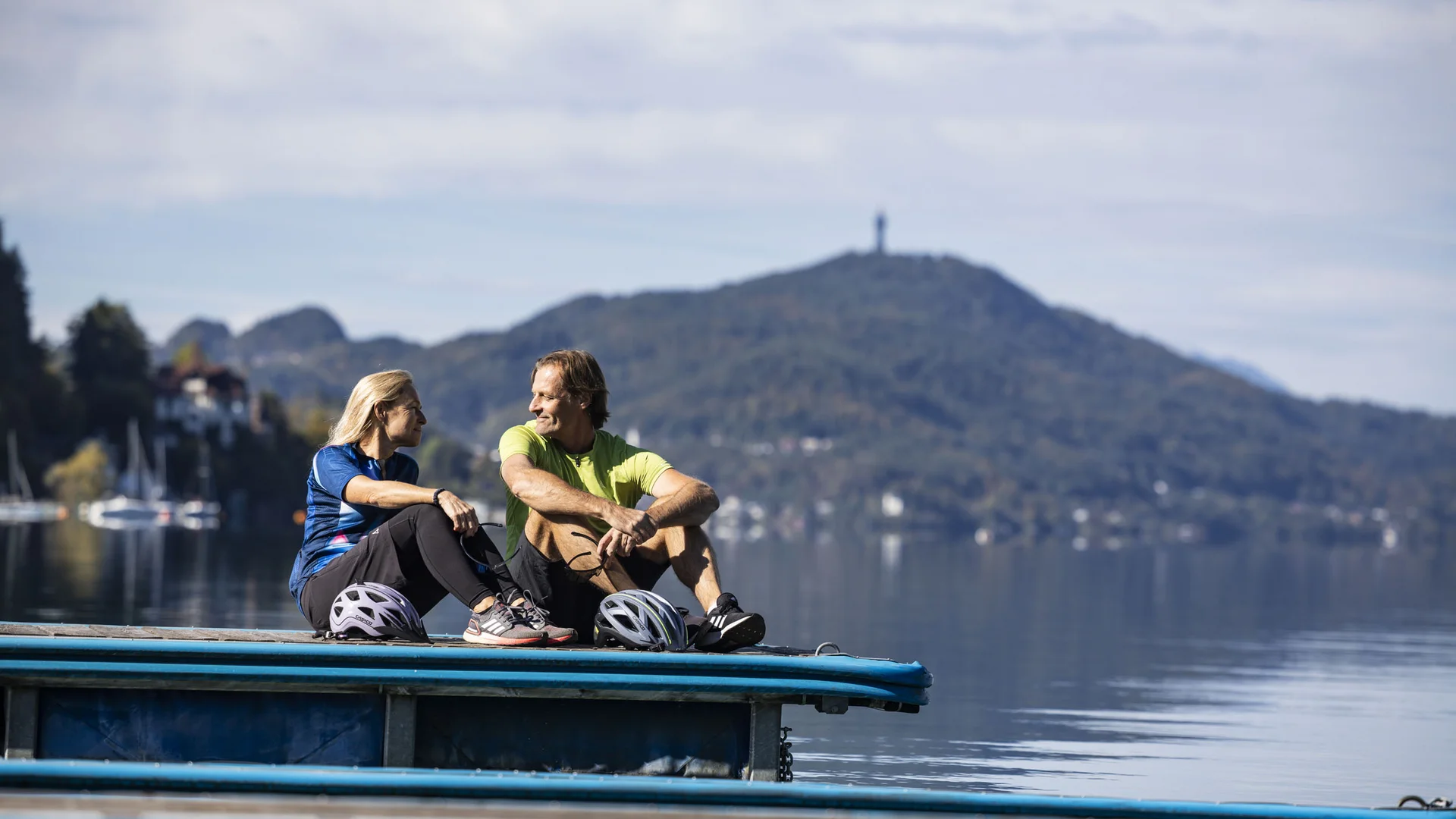 Mann und Frau mit Fahrradhelmen sitzen am Seeufer mit Berg und Wasser im Hintergrund