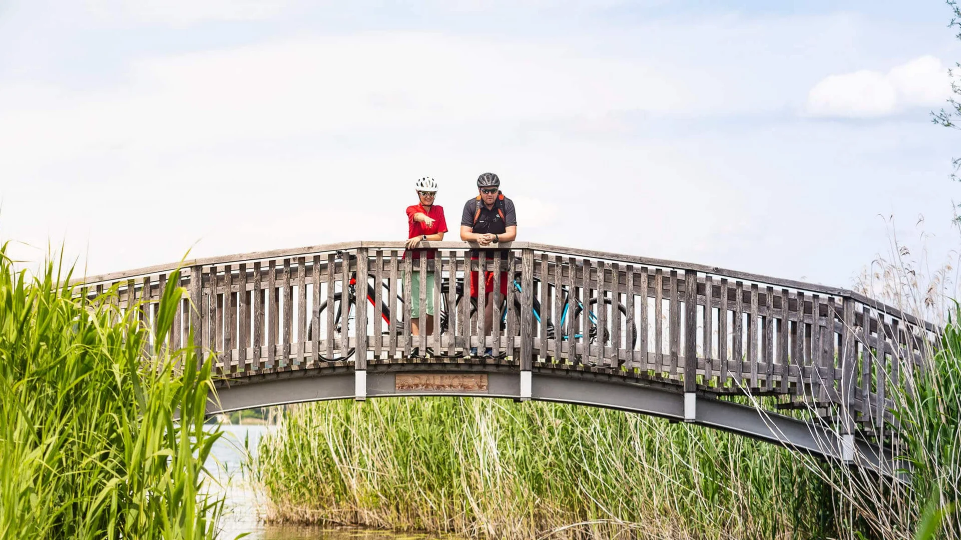 Wissenswertes © Velontour Zwei Radfahrer auf einer Holzbrücke über einen Fluss mit Schilf