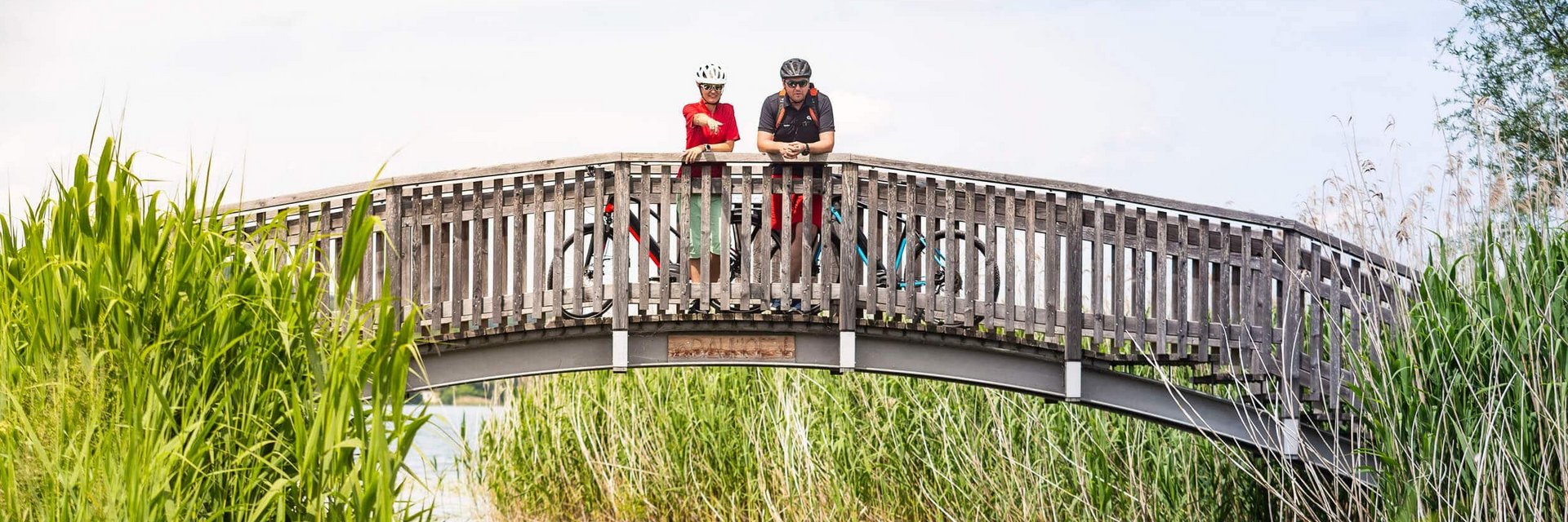 Zwei Radfahrer auf einer Holzbrücke über einen Fluss mit Schilf