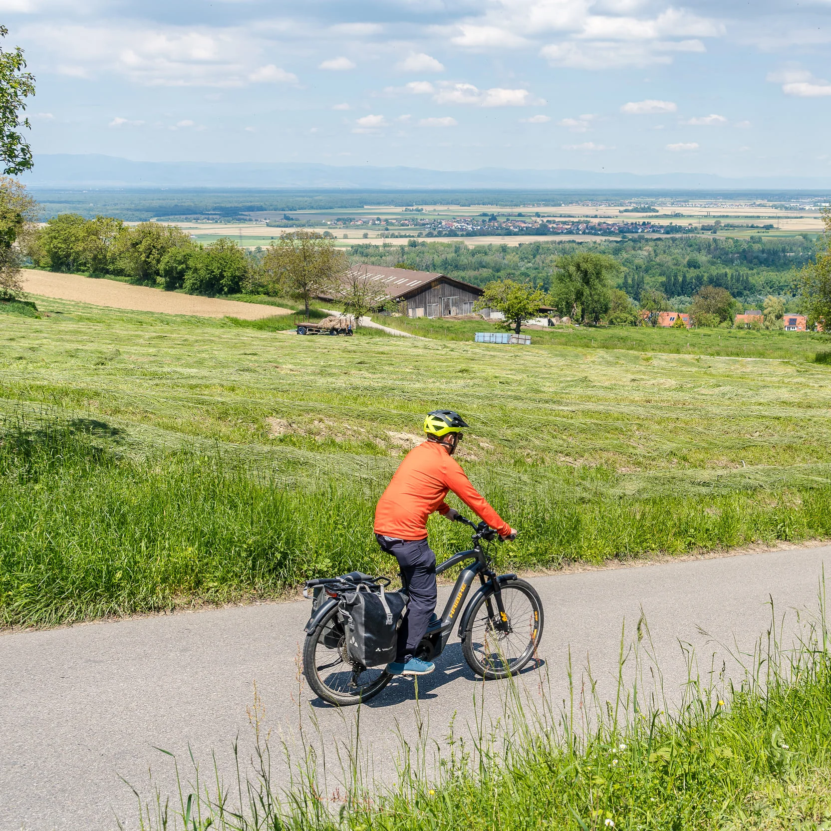 Person fährt Fahrrad auf Landstraße mit grünen Feldern und Dorf im Hintergrund