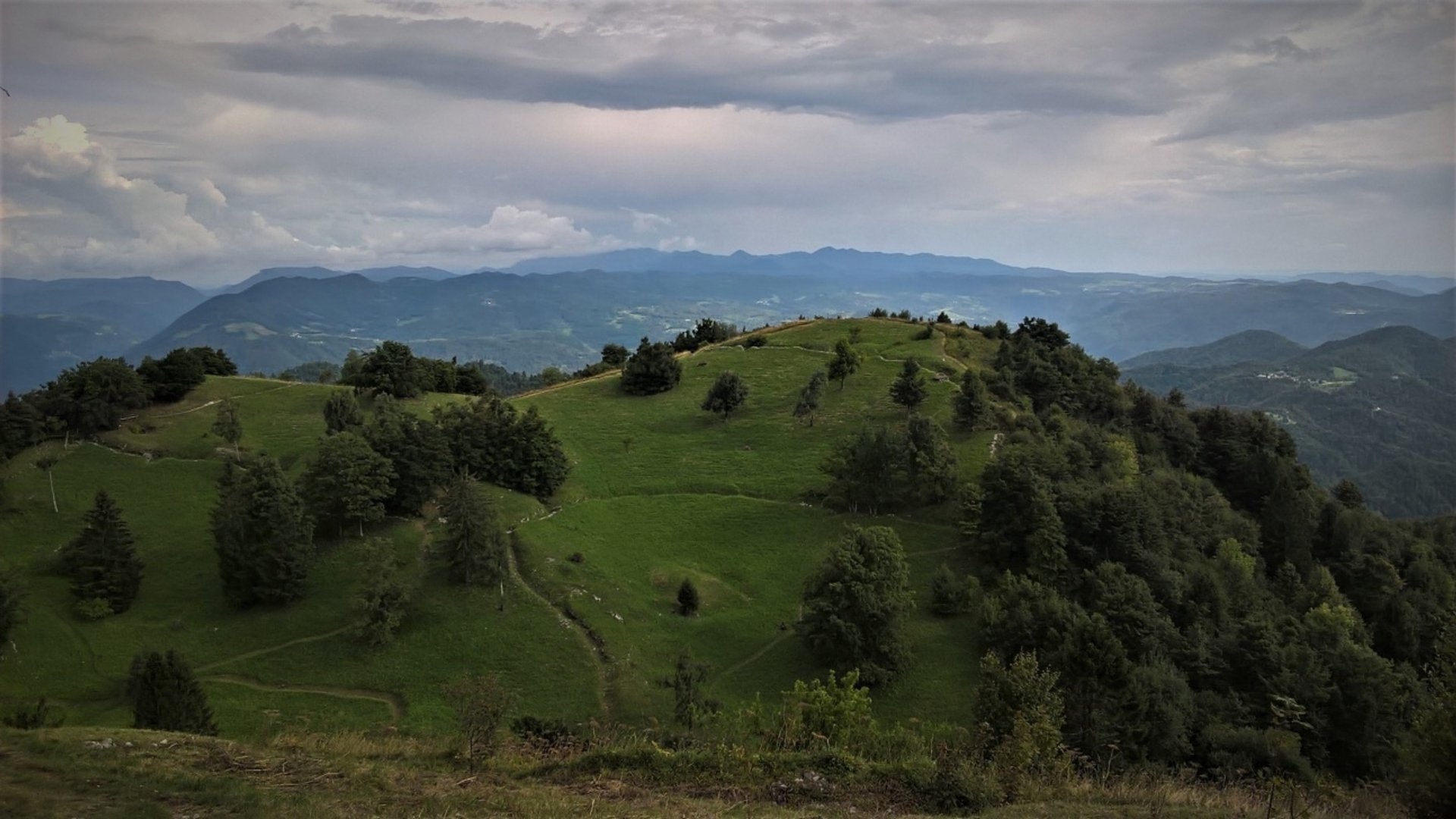 Green forested hill under cloudy sky with mountains in the background