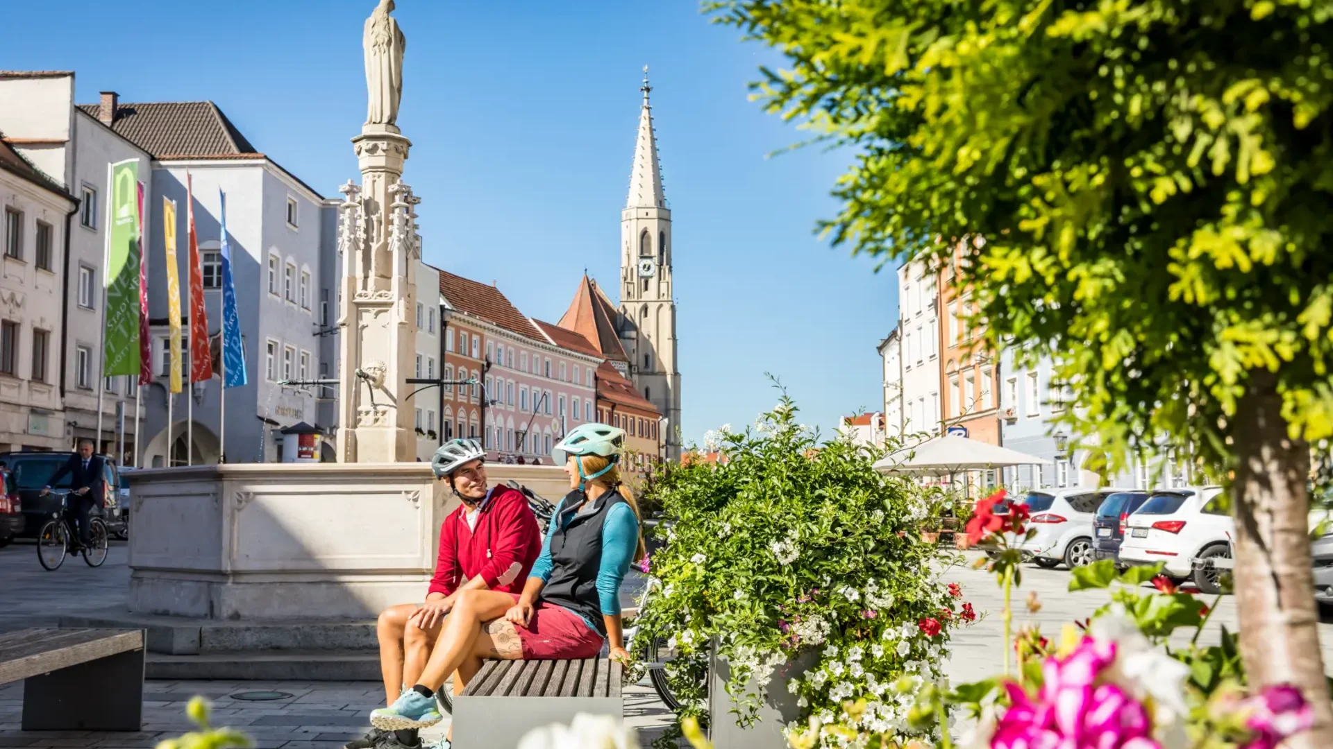Zwei Radfahrer sitzen auf einer Bank in einer sonnigen Stadt mit Kirche und Blumen