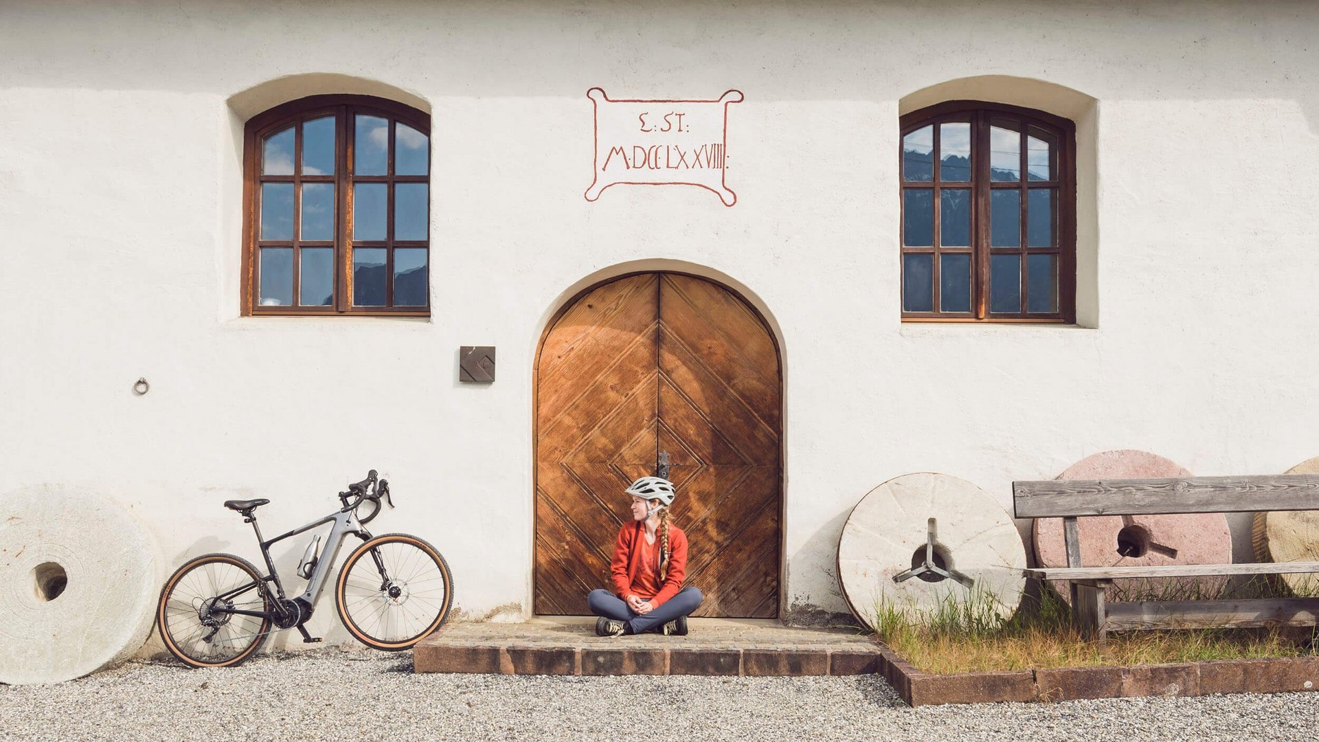 Woman with helmet sitting by wooden door of house with bike and millstones