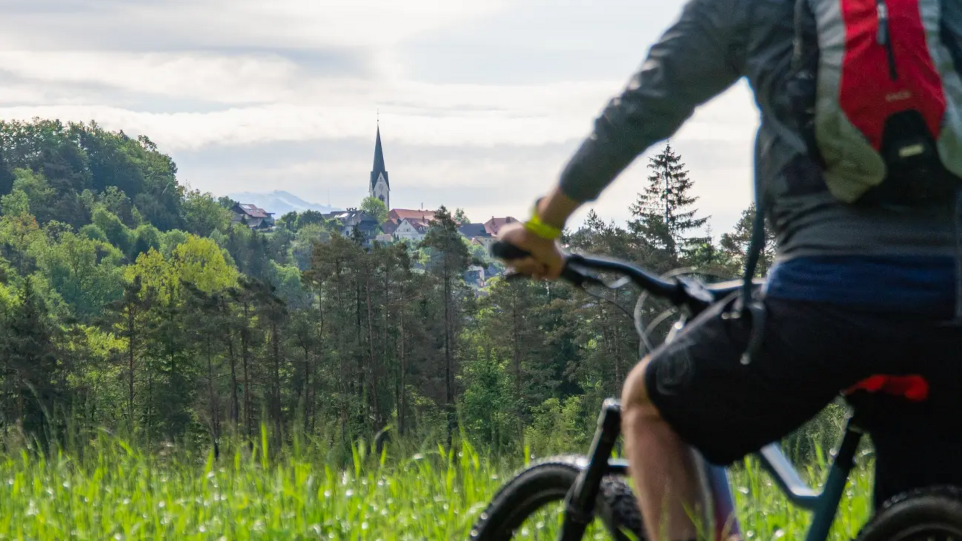 Cyclist with red backpack on mountain bike in green field near village church
