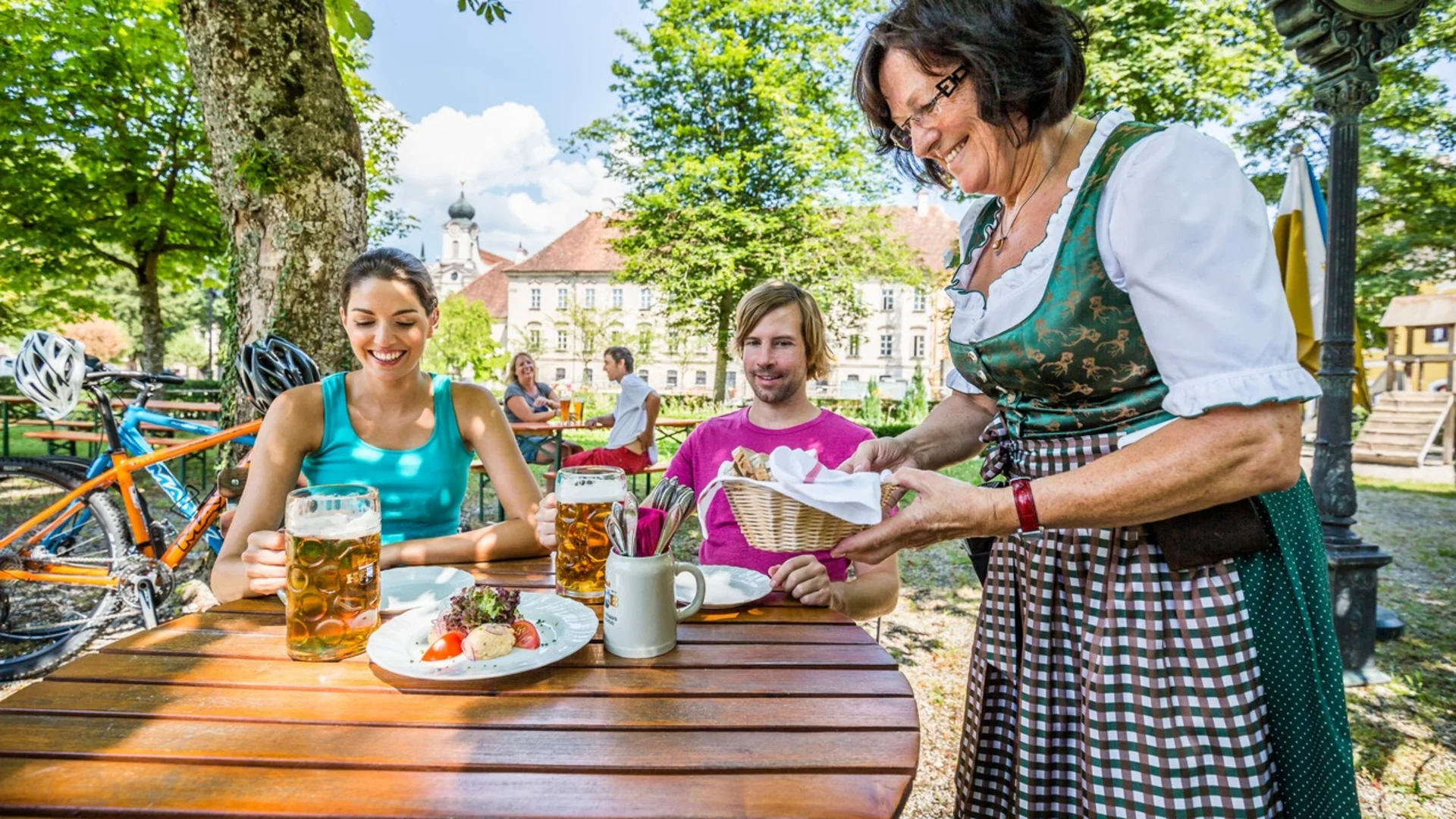 Frau in Tracht serviert Brot an Gäste im Biergarten an einem sonnigen Tag