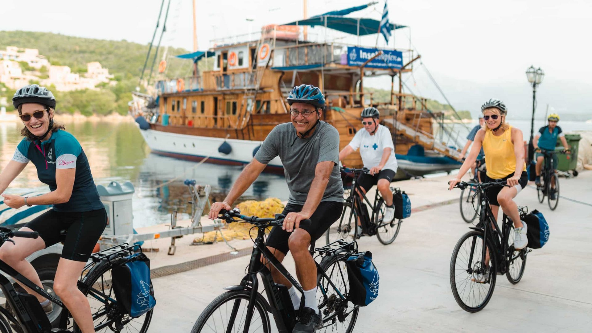 Group of cyclists wearing helmets riding along a dock with a wooden boat nearby