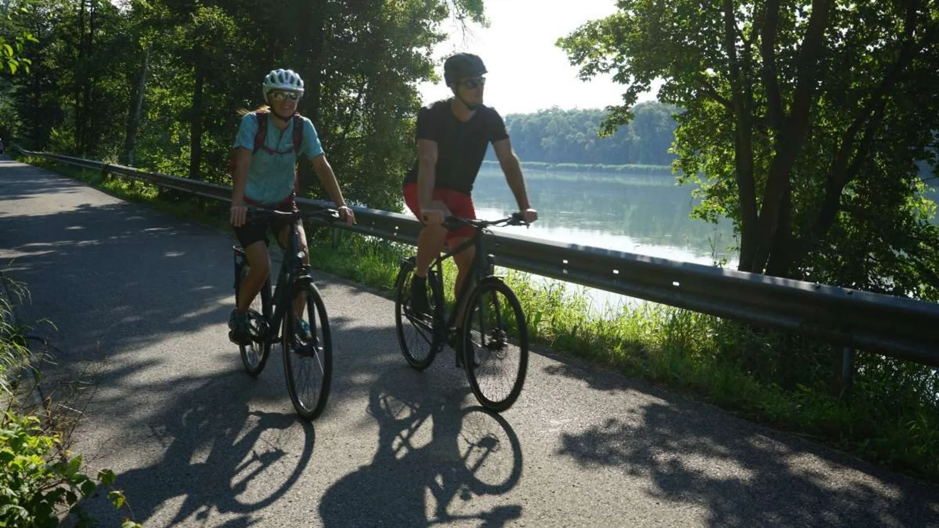 Zwei Radfahrer fahren auf einem Weg neben einem See bei sonnigem Wetter