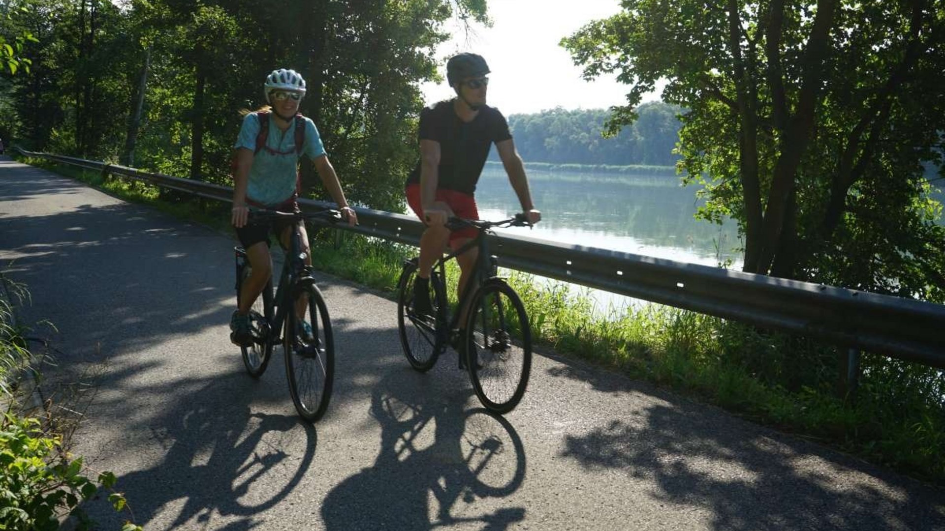 Two cyclists riding on a path beside a lake on a sunny day