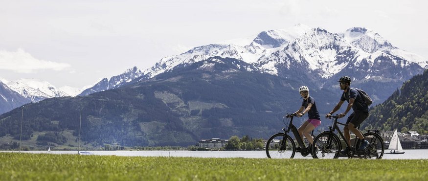 Two cyclists riding bikes near a lake with snow-covered mountains