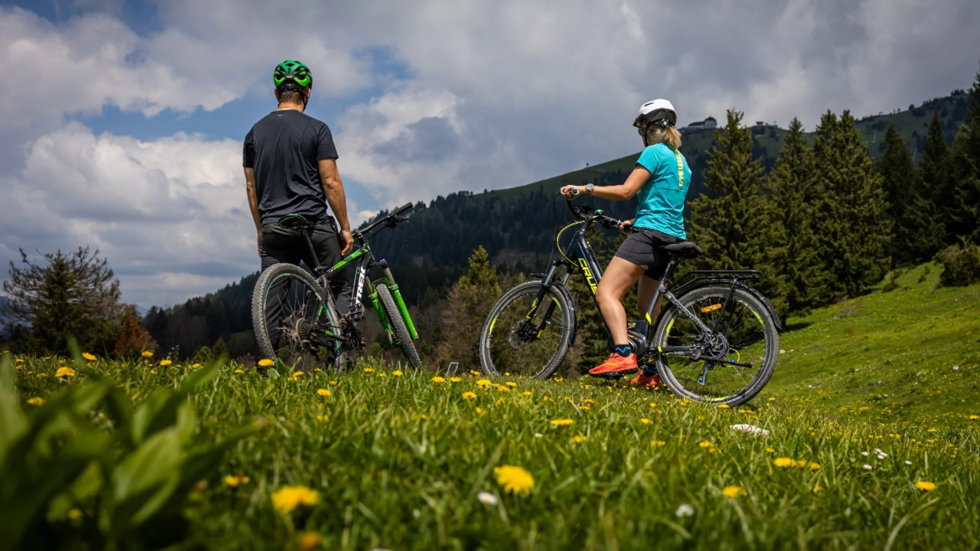 Twee fietsers op bloeiende weide met bosrijke berg onder bewolkte lucht