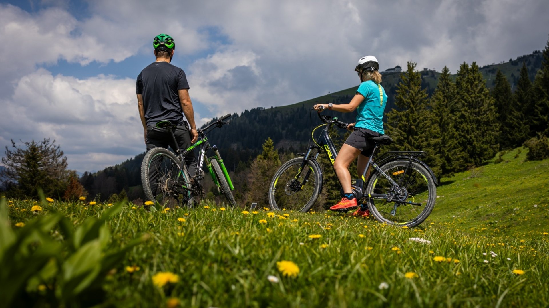 Two cyclists on a flowered meadow facing forested mountain under cloudy sky