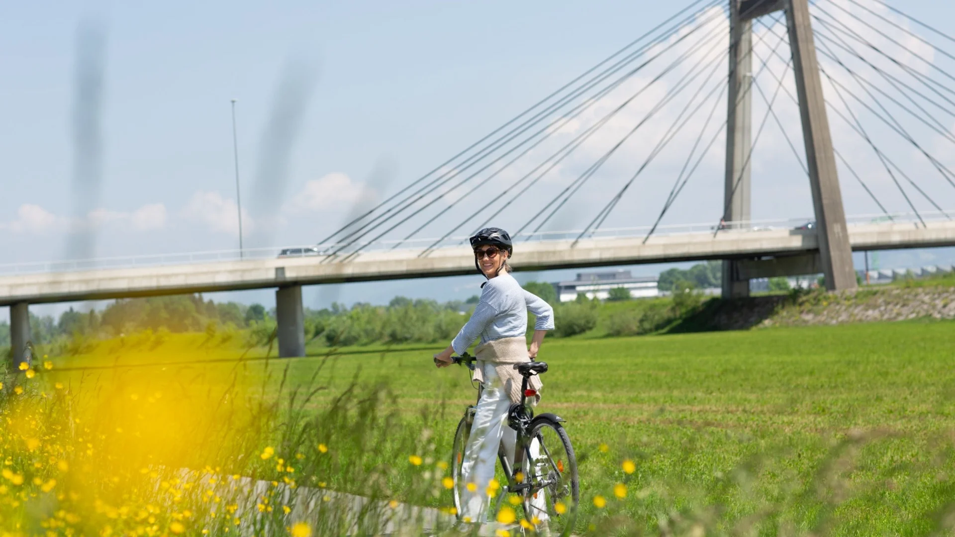 Frau mit Helm auf Fahrrad vor einer modernen Hängebrücke und grüner Wiese