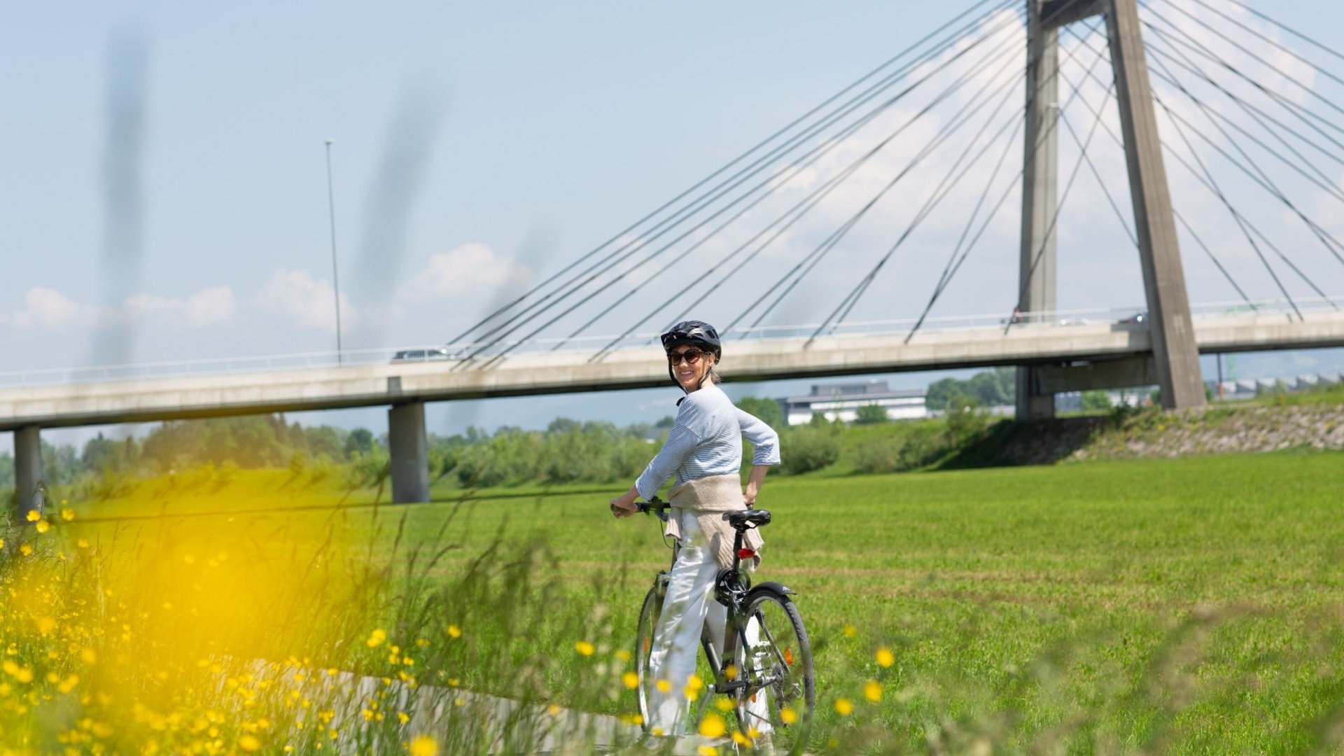 Woman wearing helmet on bicycle near a cable-stayed bridge and green field