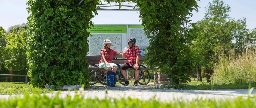 Zwei Radfahrer machen eine Pause auf einer Bank unter einem grünen Pflanzenbogen