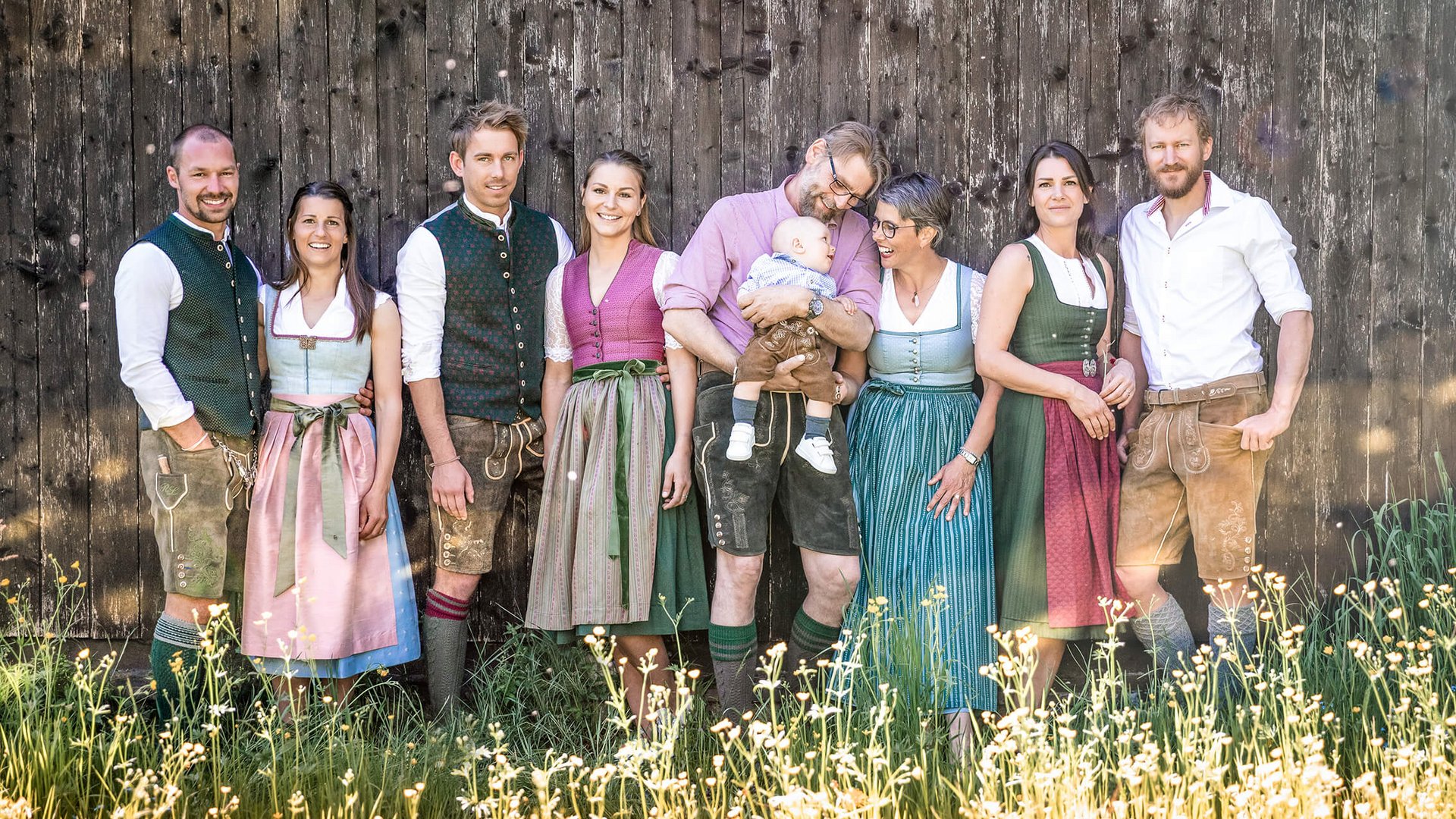 Nine people wearing traditional Bavarian clothing standing by wooden wall
