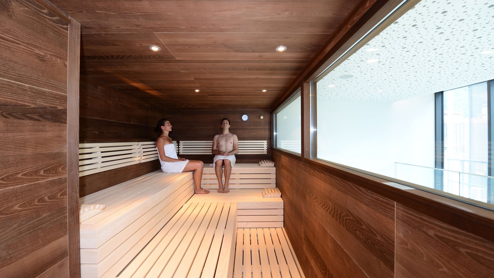 Two people sitting in a modern wooden sauna with a large window.