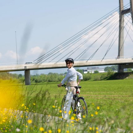 Frau mit Helm fährt Fahrrad vor einer Brücke in grüner Landschaft