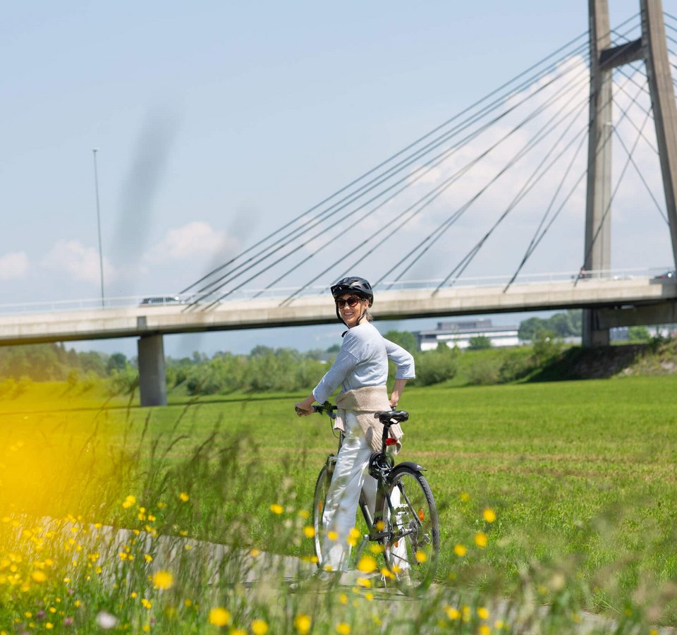 Frau mit Helm fährt Fahrrad vor einer Brücke in grüner Landschaft