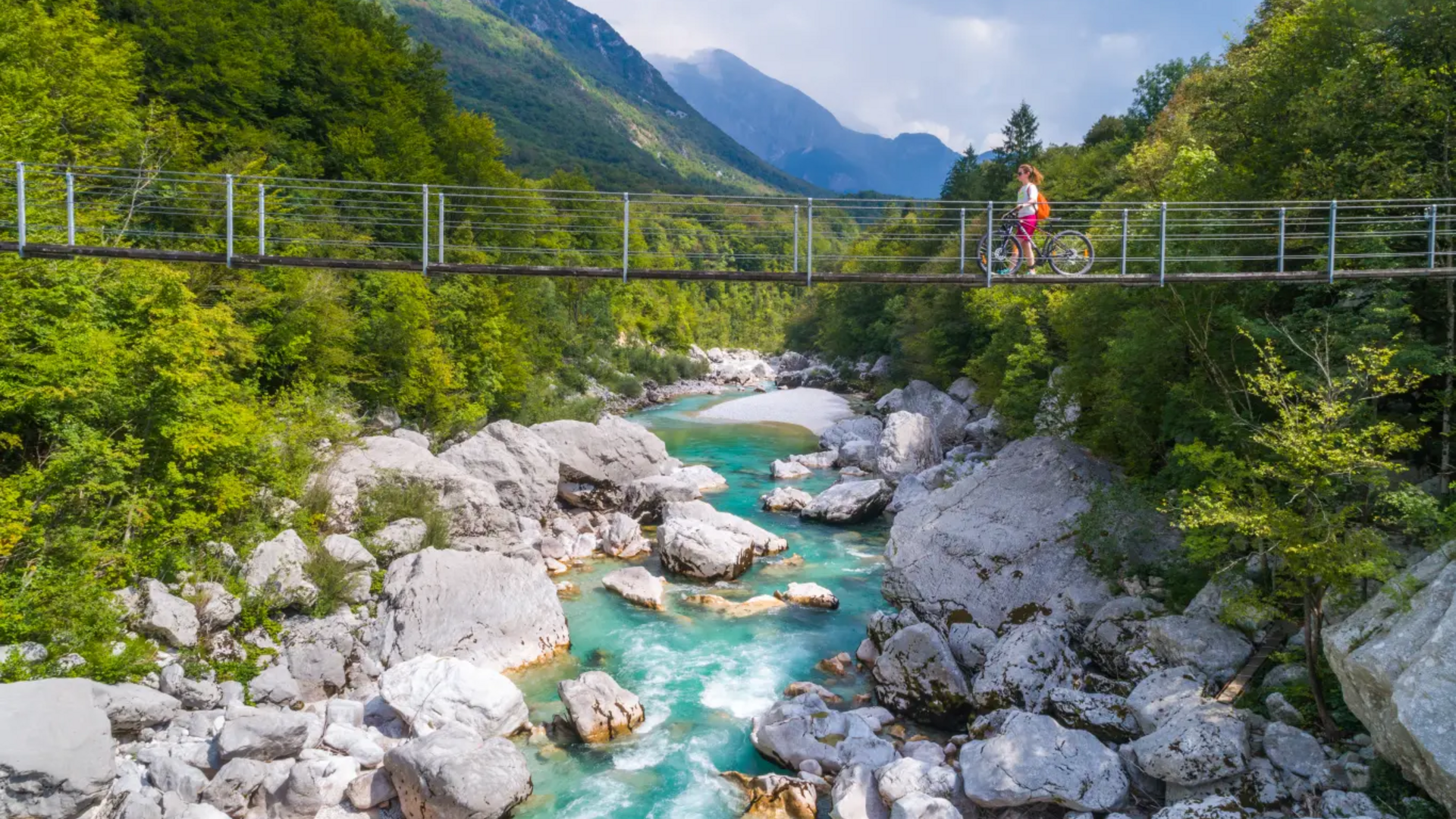 Cyclist on suspension bridge over turquoise river in mountainous landscape
