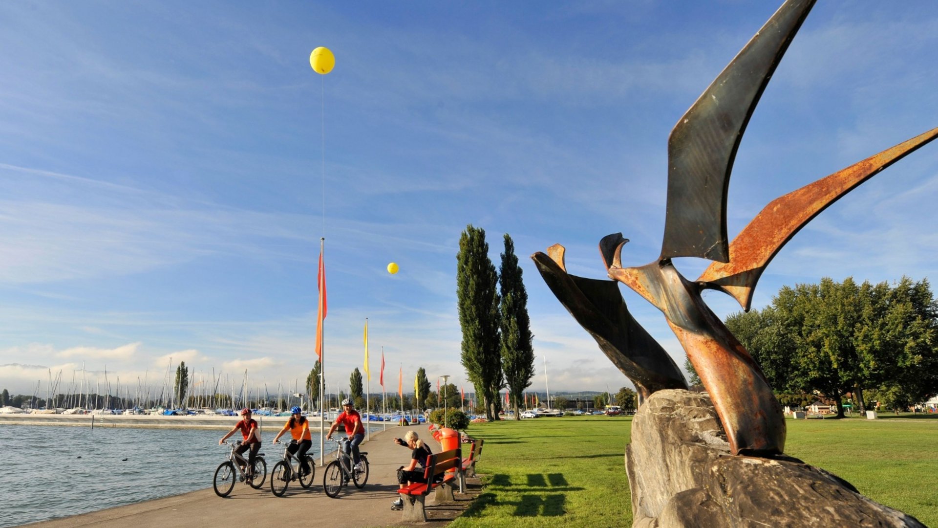 People cycling along a lake shore with sculpture and boats in the background