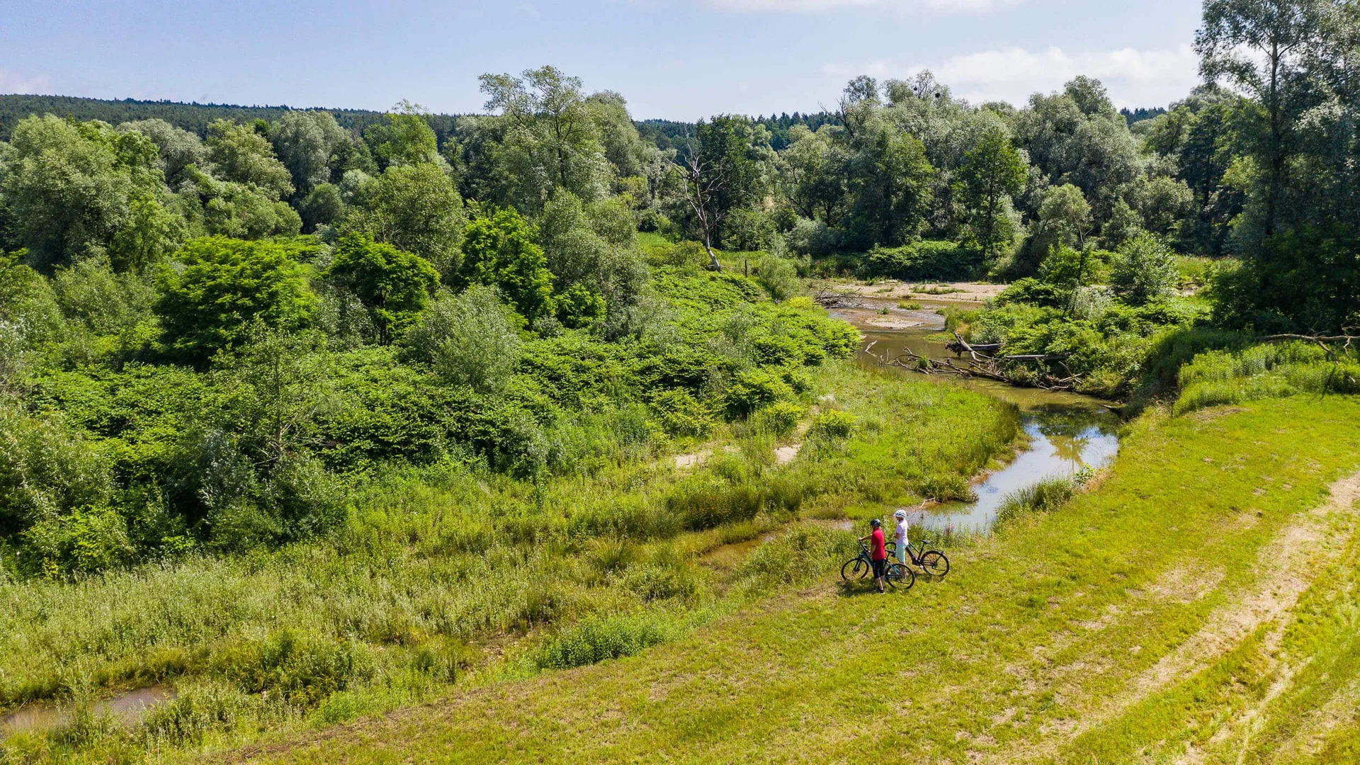 Zwei Radfahrer stehen an einem kleinen Fluss in einer grünen Naturlandschaft