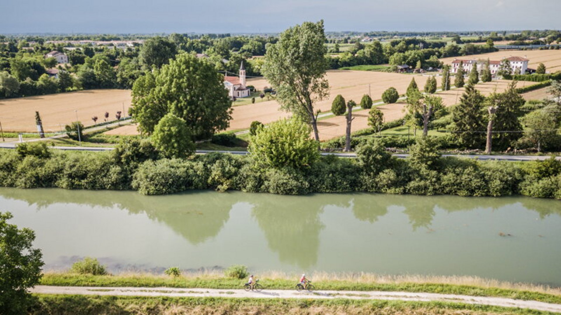 Riverbank with bike path and cyclists, fields and trees in the background