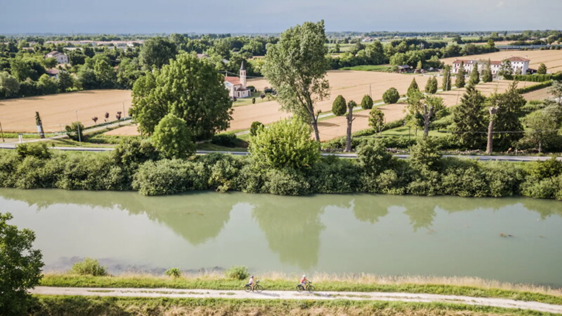 Flussufer mit Radweg und Radfahrern, Felder und Bäume im Hintergrund