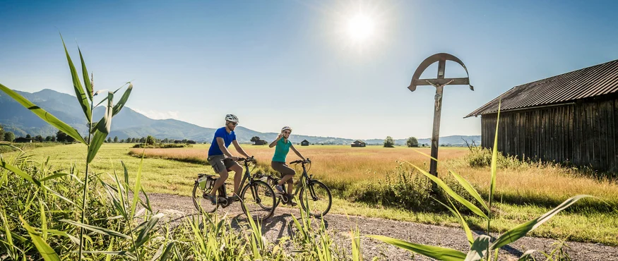 Im Sattel durchs Tölzer Land entlang von Isar & Loisach © Tourist Information Kochel am See - Thomas Kujat Zwei Radfahrer auf Landweg bei sonnigem Wetter vor Berglandschaft