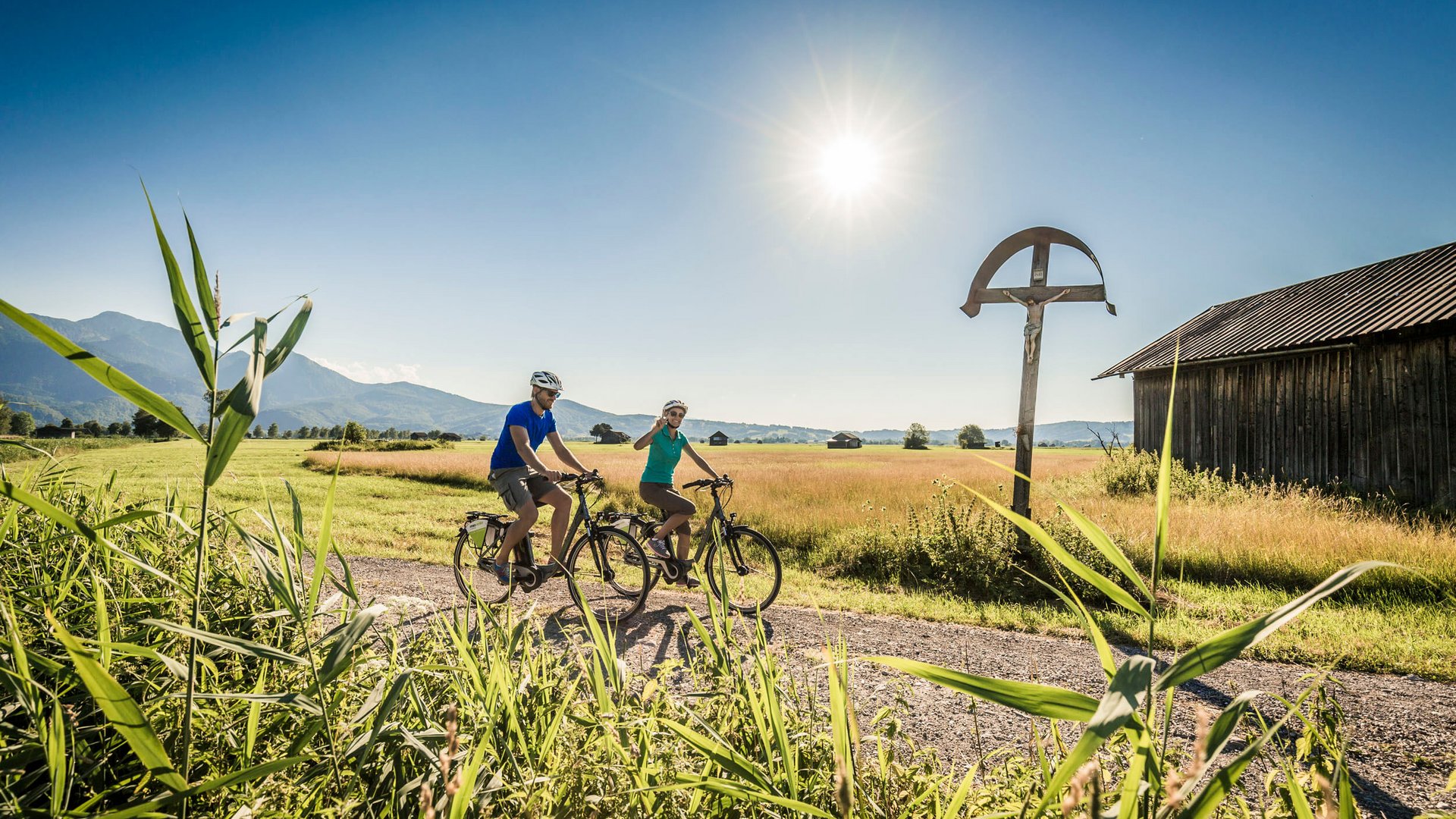 Two cyclists on country path under bright sun with mountains in background