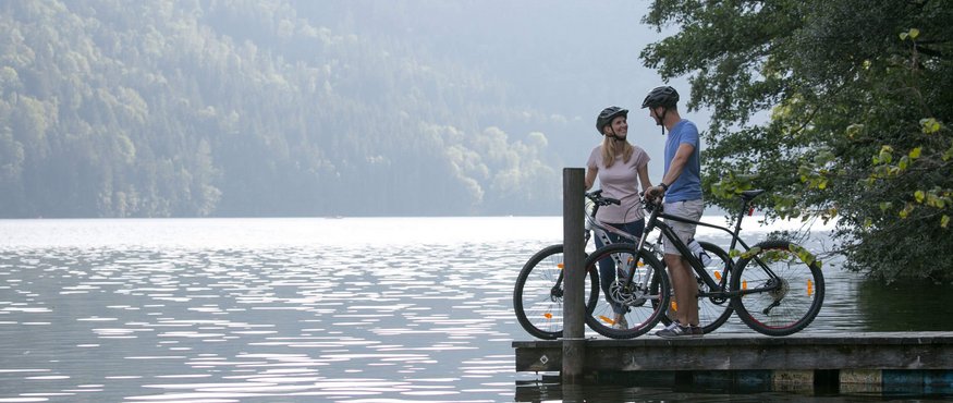 Two cyclists with bikes on a pier by a lake surrounded by forest