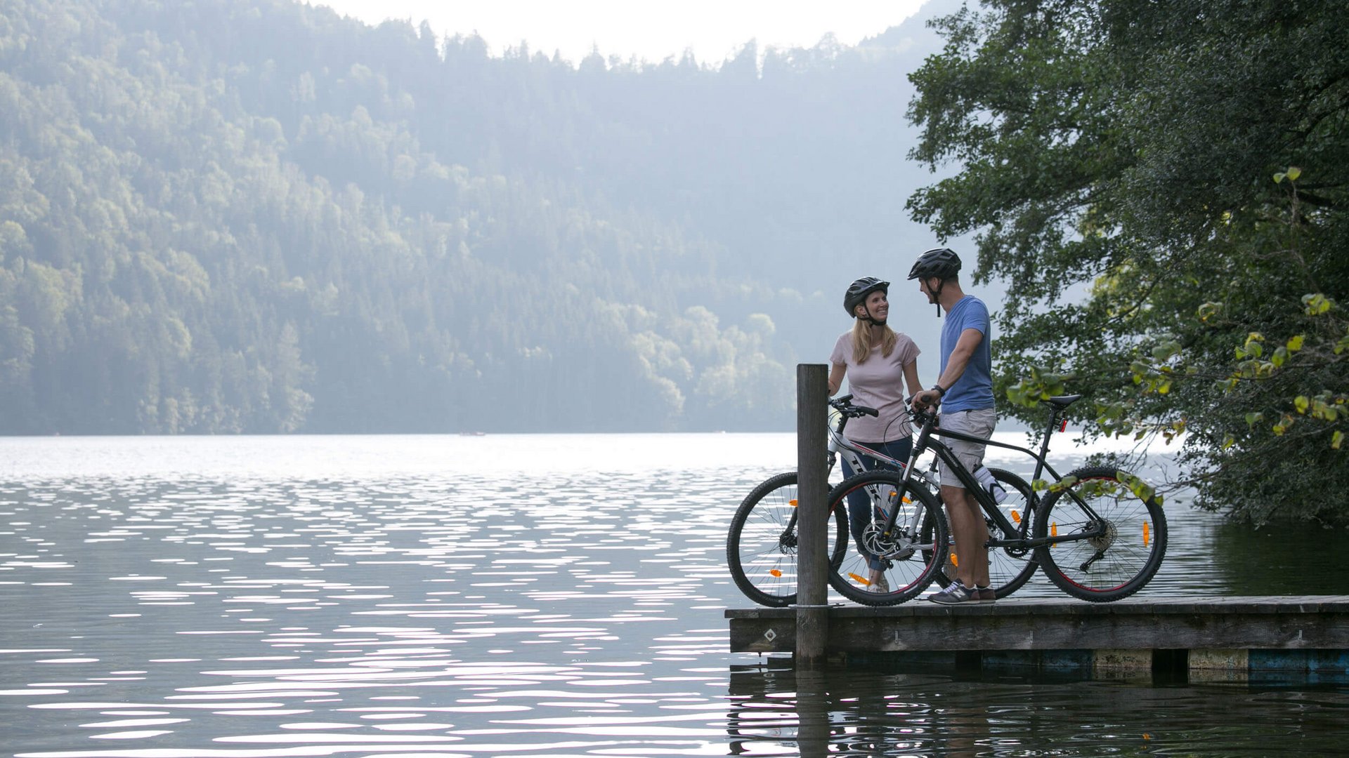 Two cyclists with bikes on a pier by a lake surrounded by forest