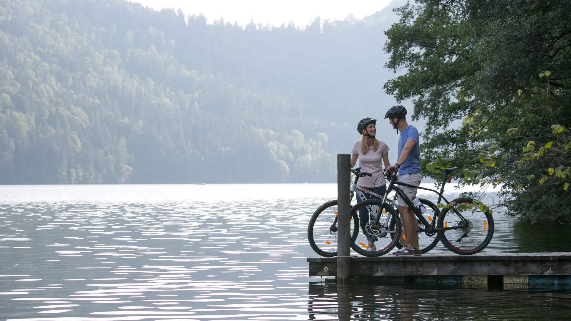 Zwei Radfahrer auf Steg am See mit Wald im Hintergrund