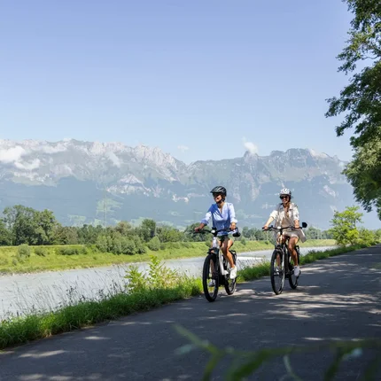 Twee vrouwen fietsen langs een rivier met bergen op de achtergrond