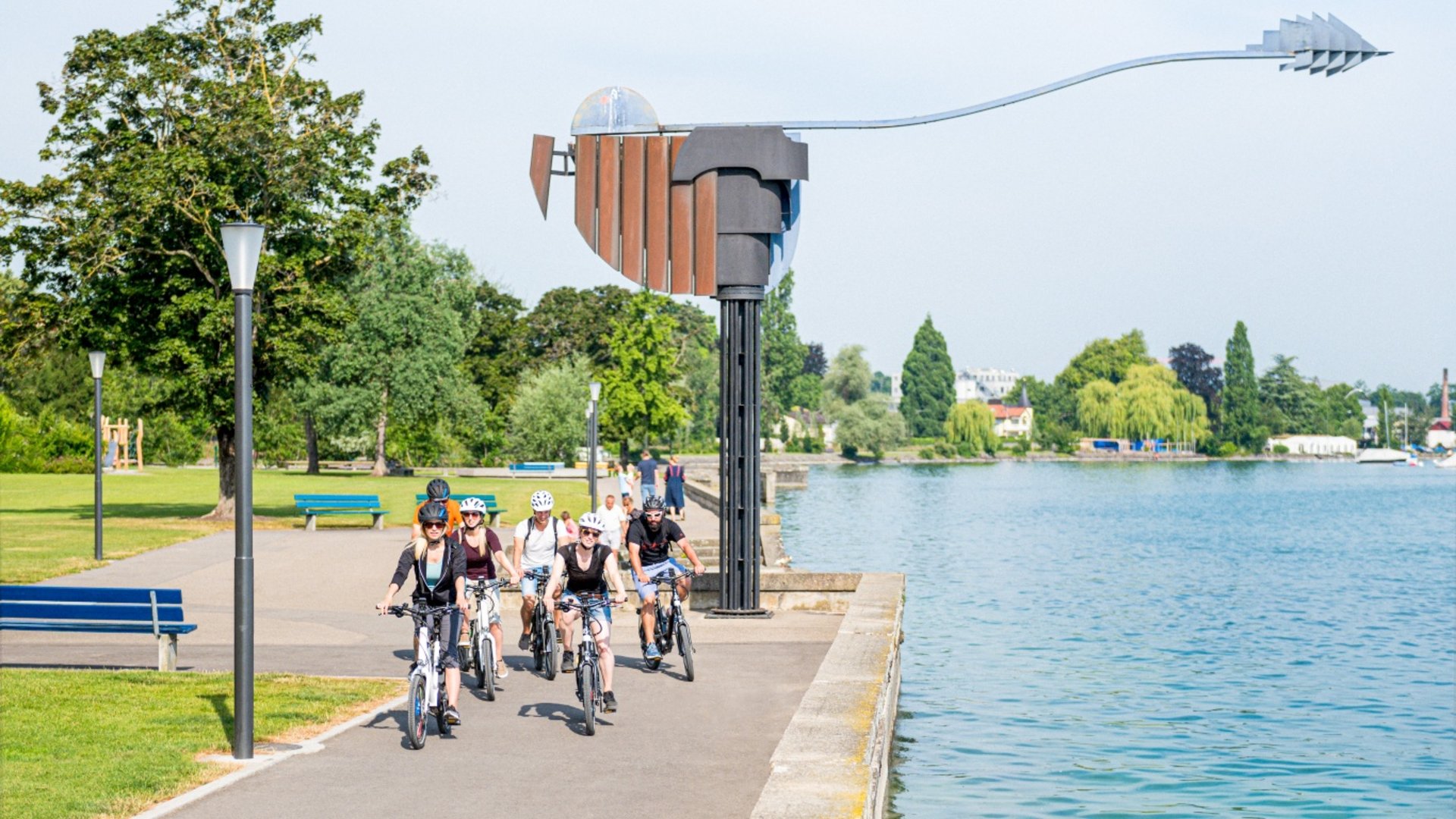 Group of cyclists on a lakeside path with a sculpture in the background
