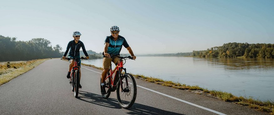 Two people cycling on a path by a river on a sunny day