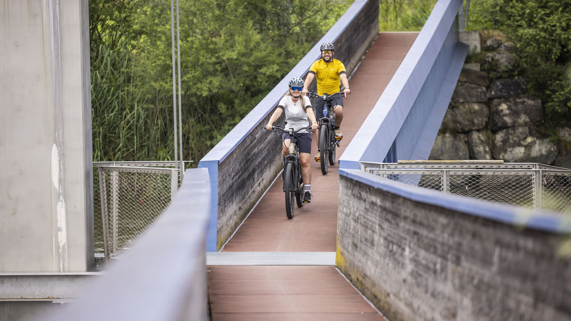 Two cyclists riding on a narrow bridge surrounded by trees