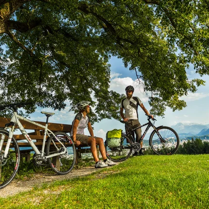 Anthering: Radurlaub zwischen Moor & Mozartstadt © Markus Greber Zwei Radfahrer machen Pause auf einer Bank unter einem Baum mit Bergblick