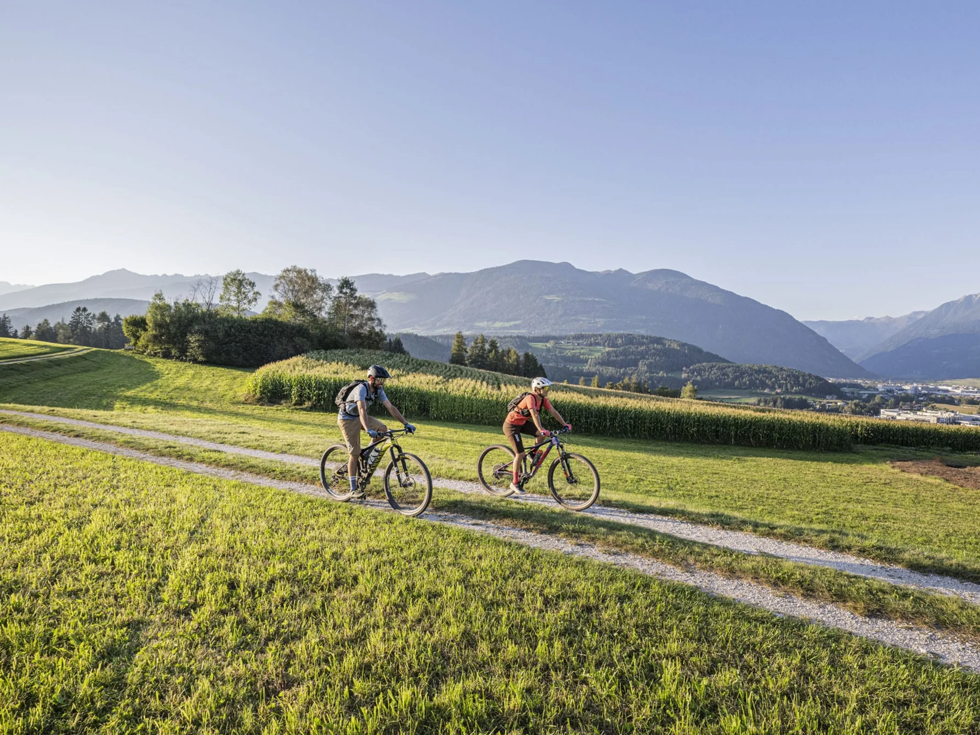 Zwei Radfahrer auf einem Weg durch grüne Wiesen mit Bergen im Hintergrund