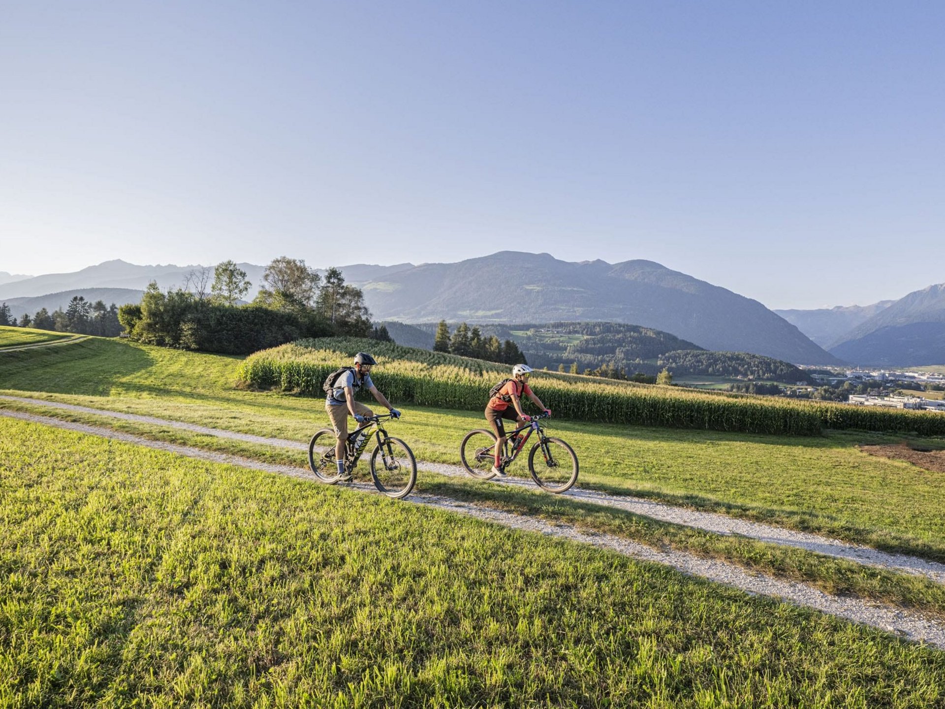 Zwei Radfahrer auf einem Weg durch grüne Wiesen mit Bergen im Hintergrund