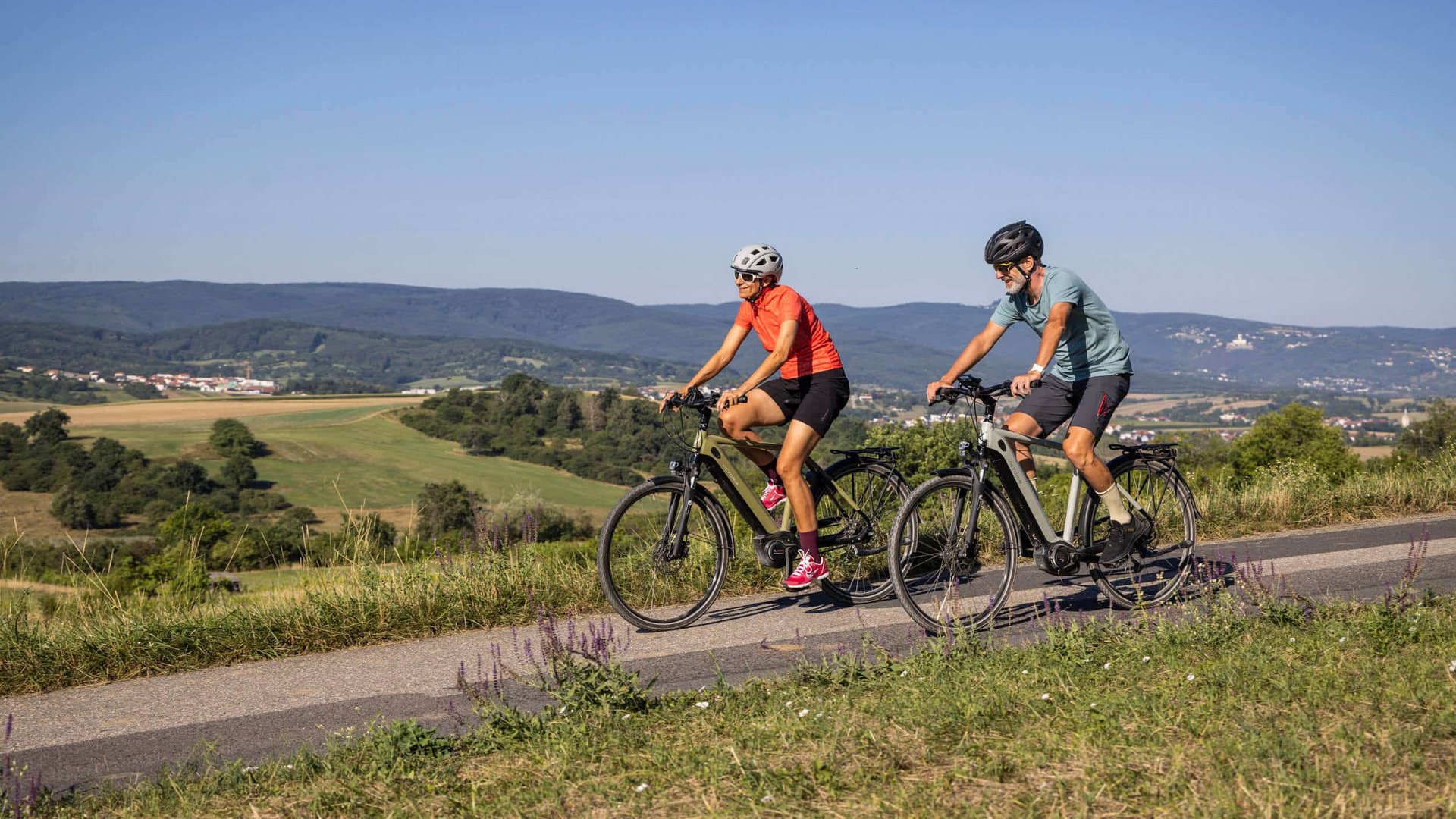 Two cyclists riding on a country road through hilly landscape under clear sky