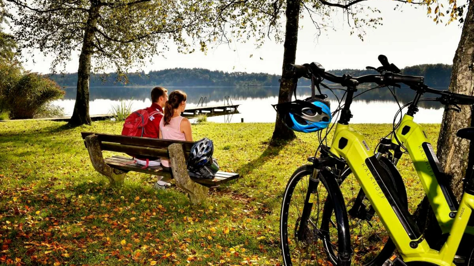 Paar sitzt auf Bank am See, zwei gelbe Fahrräder stehen an Bäumen