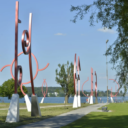 Red metal art sculptures lining a lakeside path under clear sky