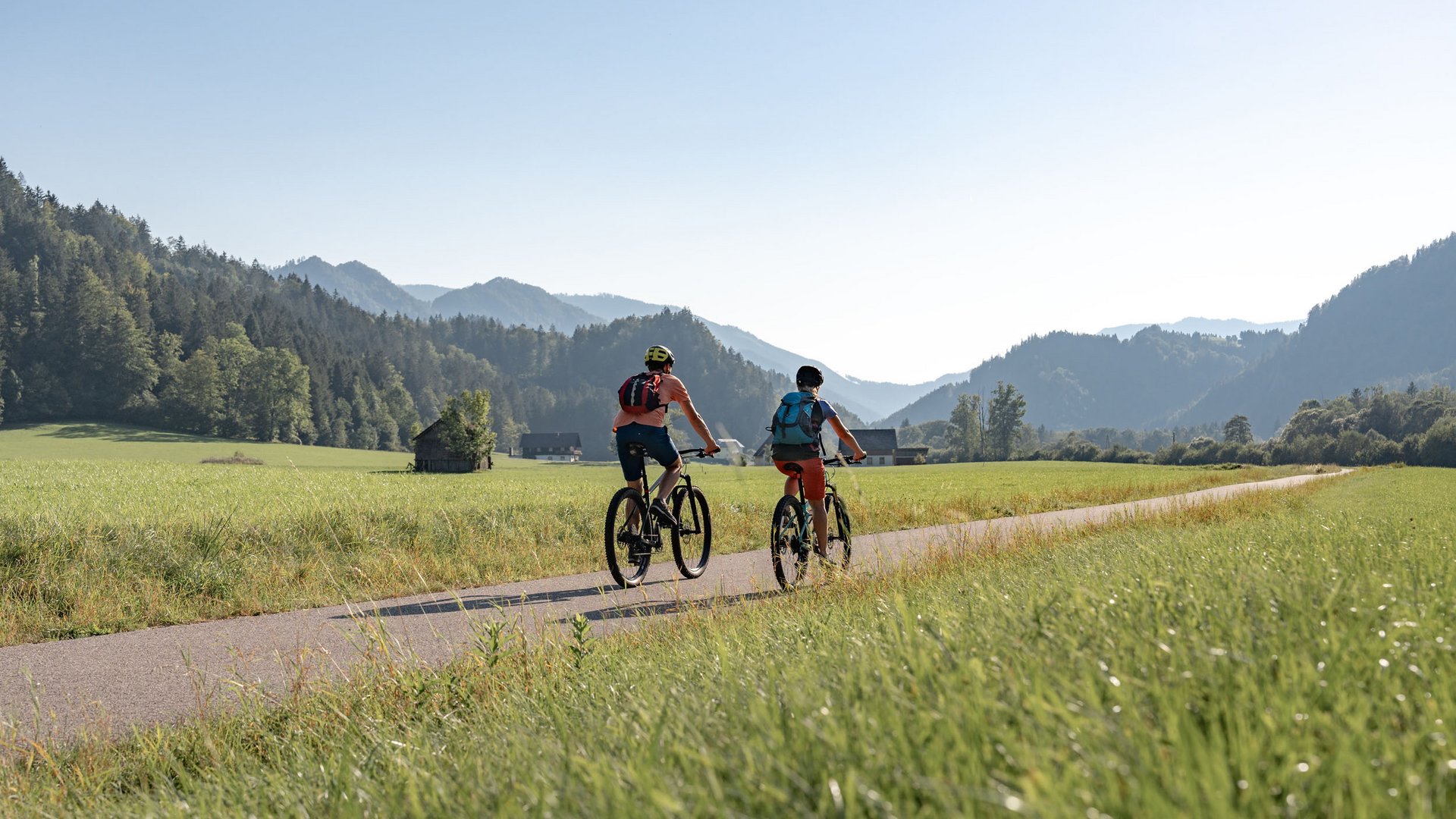 Two cyclists riding on a country road with mountains and green fields
