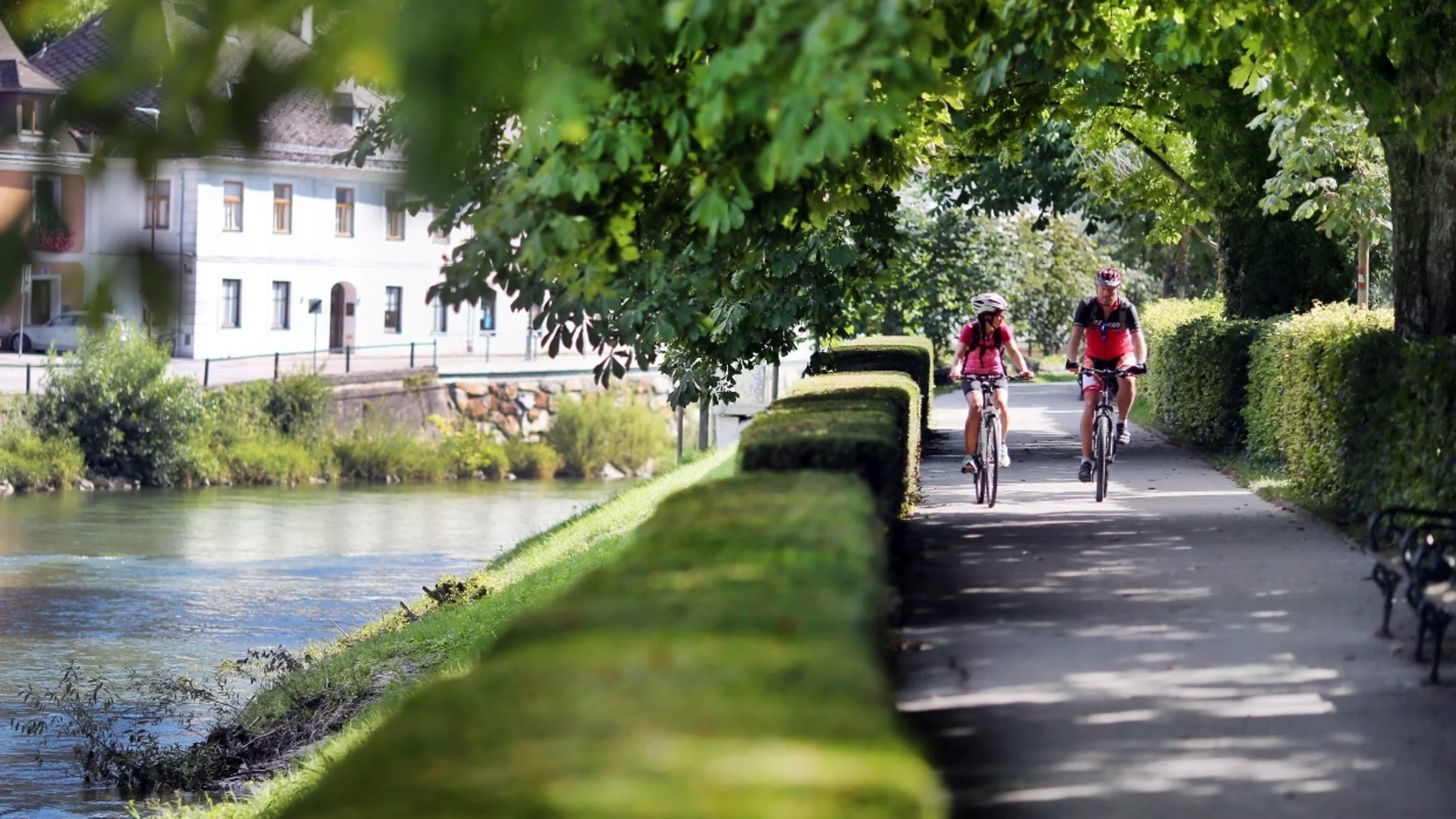Tour-Detail Zwei Radfahrer fahren an einem Fluss und Bäumen entlang in einer sonnigen Landschaft.