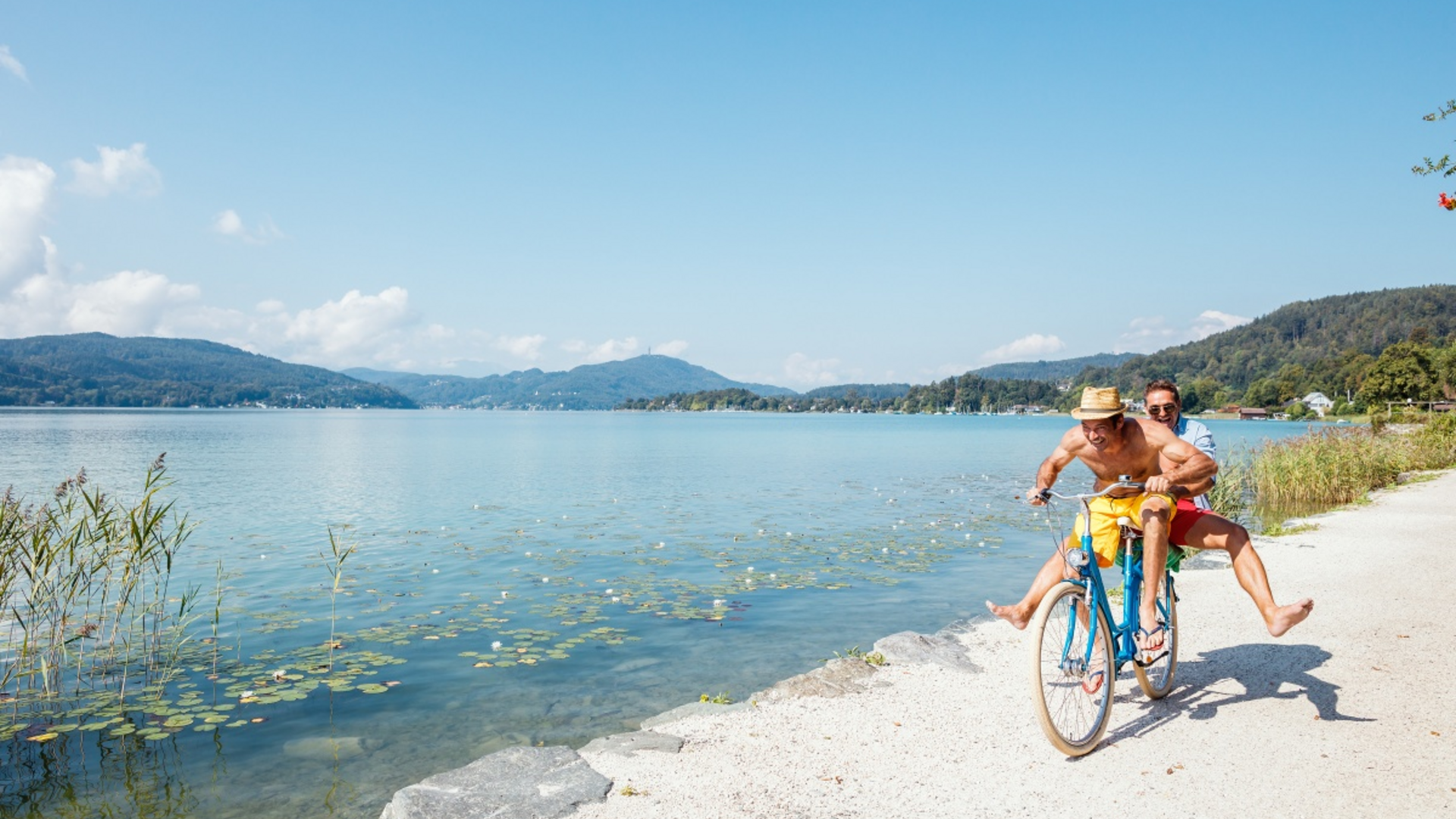 Two men happily riding a bicycle by the lakeside on a sunny day
